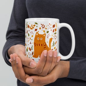 May include: White ceramic mug with a cartoon orange cat wearing a scarf and surrounded by autumn leaves and flowers.