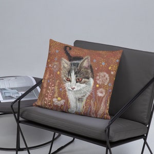 May include: A throw pillow with a brown background featuring a white and gray tabby cat walking through a field of flowers. The cat has green eyes and a white chest.