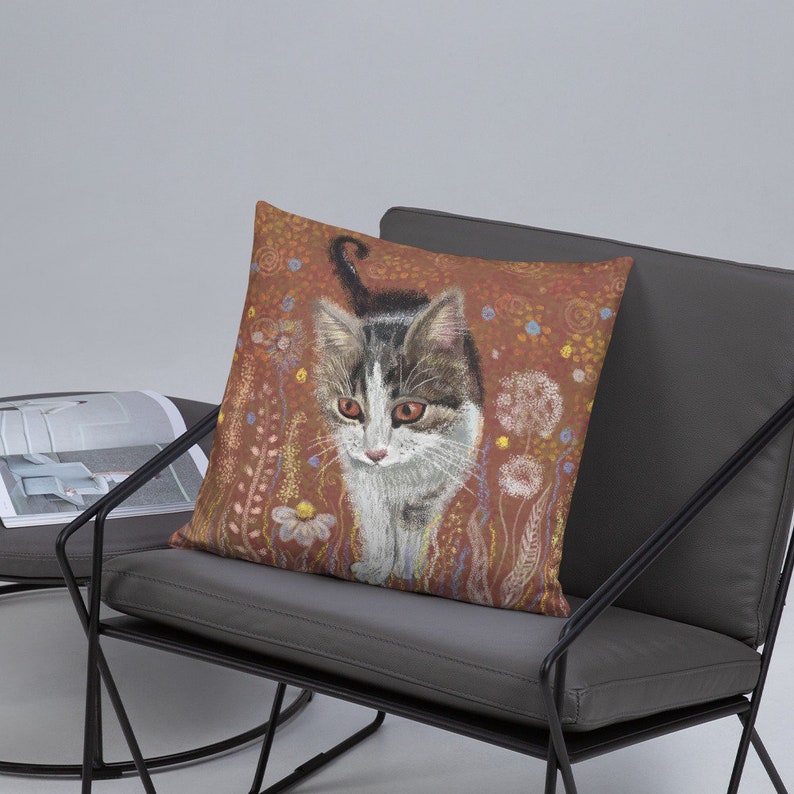 May include: A throw pillow with a brown background featuring a white and gray tabby cat walking through a field of flowers. The cat has green eyes and a white chest.