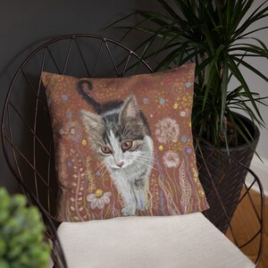 May include: A throw pillow with a colorful illustration of a cat with brown, white, and gray fur. The cat is walking through a field of flowers and dandelions. The background is a warm brown color.