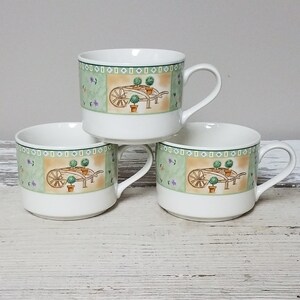 May include: Three white ceramic mugs with a green, brown and white floral design featuring a wheelbarrow with potted plants. The mugs are stacked on top of each other.