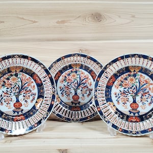 May include: Three decorative ceramic plates with a blue and white lattice design and a floral pattern in shades of red, orange, and gold. The plates are displayed on a wooden surface.