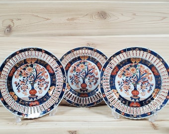 May include: Three decorative ceramic plates with a blue and white lattice design and a floral pattern in shades of red, orange, and gold. The plates are displayed on a wooden surface.