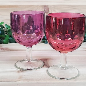 May include: Two clear glass wine goblets with a pink, iridescent finish. The goblets have a round bowl and a long stem.