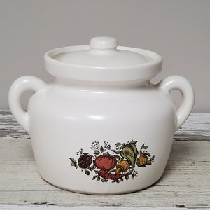 May include: White ceramic jar with a lid and two handles. The jar has a floral design with vegetables, including a tomato, pepper, mushroom, and carrot.