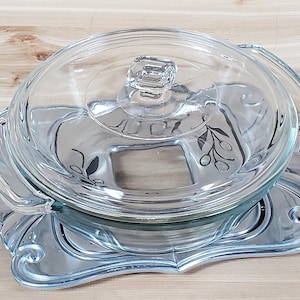 May include: A clear glass baking dish with a lid and two handles. The dish has a floral design on the bottom. The dish is sitting on a silver-colored trivet.