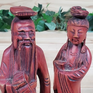May include: Two intricately carved wooden figurines, one male and one female, with detailed facial features and traditional clothing. The figurines are painted in a rich reddish-brown colour.