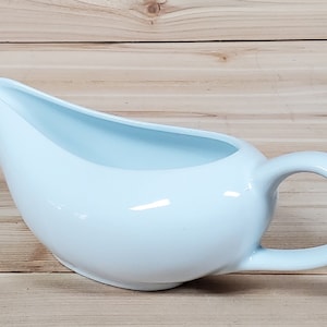 May include: A light blue ceramic gravy boat with a handle. The boat is smooth and glossy.