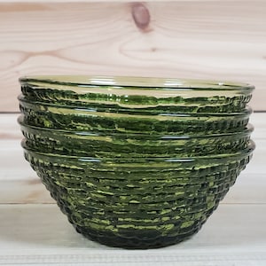 May include: A stack of four green glass bowls with a textured, woven pattern. The bowls are a deep, forest green color and have a rounded shape. The bowls are stacked on top of each other, with the top bowl slightly visible.