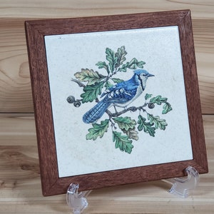 May include: A blue jay bird perched on a branch with green leaves. The bird is painted on a white ceramic tile with a brown wooden frame. The tile is on a stand.