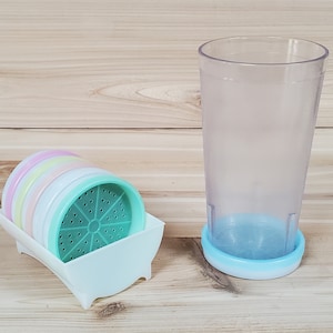 May include: A set of colorful plastic coasters with a green coaster with holes in the center. The coasters are in a white plastic holder. A clear plastic cup with a blue plastic ring around the bottom is next to the coasters.