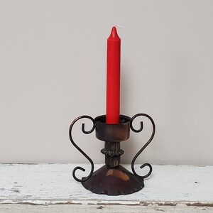 May include: A red candle sits in a decorative metal candle holder with a scrolled design. The holder is made of dark metal with a copper finish.