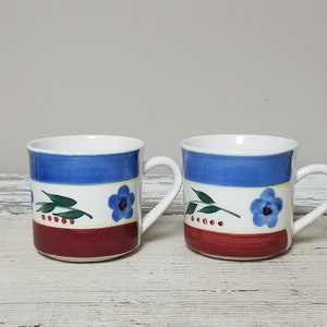 May include: Two white ceramic mugs with blue, red, yellow and green floral designs. The mugs have a white background with a blue stripe, a yellow stripe and a red stripe. The floral design features a blue flower with a green stem and leaves.