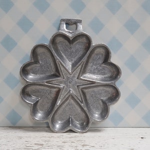 May include: A vintage aluminum heart-shaped waffle iron with a star in the center. The iron has a handle on top and is designed to make six heart-shaped waffles.