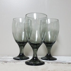 May include: Three gray glass wine goblets with a dark gray base. The goblets are clear and have a slight smoky tint.