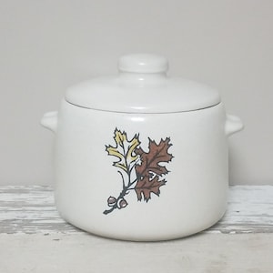 May include: White ceramic soup pot with a lid, featuring a brown and yellow leaf design. The pot has two handles and a knob on the lid.