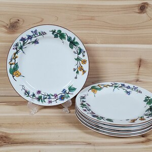 May include: A set of six white ceramic dinner plates with a floral design featuring purple, yellow, and green flowers. The plates have a brown rim.