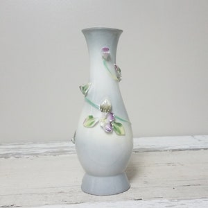 May include: A small, white ceramic vase with a blue glaze and 3D floral accents. The vase features delicate purple flowers and green leaves.
