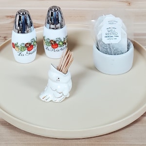 May include: A beige round tray with a white ceramic salt and pepper shaker set, a white ceramic toothpick holder shaped like a bird, and a white ceramic container with a tea bag inside. The salt and pepper shakers have a floral design with the words "La Salt" and "Le Poivre" on them. The tea bag is labeled "Well Rested Herbal Tea."