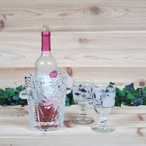 May include: A crystal wine cooler with a bottle of white wine inside, and two clear wine glasses with a floral design on the side. The glasses and cooler are on a wooden surface with green ivy.