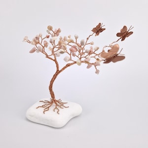 Könnte beinhalten: Handgefertigte Kupferdraht-Baumskulptur mit rosa und weißen Edelsteinblättern und Kupfer-Schmetterlingen. Der Baum steht auf einem weißen Steinsockel, ein einzigartiges Dekorationsstück.