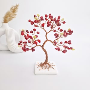 Peut inclure: Une sculpture décorative en forme d'arbre en fil de cuivre, avec un tronc et des branches cuivrées. Les feuilles de l'arbre sont faites de pierres précieuses rouges et jaunes. L'arbre repose sur une base carrée blanche.