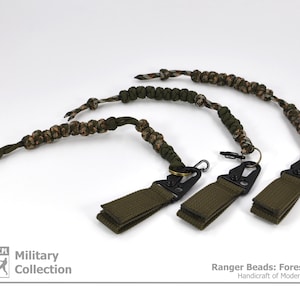 Può includere: Tre portachiavi in paracord mimetico verde e marrone con clip in metallo nero e anelli in tessuto verde. I portachiavi sono etichettati "Ranger Beads: Forest Camo Handcraft of Modern Evolution".
