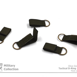 Pode incluir: Cinco adaptadores t&aacute;ticos de anel em D de cor verde oliva com an&eacute;is de lat&atilde;o. Os adaptadores s&atilde;o feitos de fita e t&ecirc;m um fecho de velcro. "Olive Green Color Tactical D-Ring Adapter Made with Passion" est&aacute; impresso em um fundo branco abaixo dos adaptadores.
