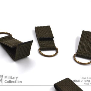 Pode incluir: Tr&ecirc;s adaptadores t&aacute;ticos de anel em D de cor verde oliva com an&eacute;is de lat&atilde;o. Os adaptadores s&atilde;o feitos de fita e t&ecirc;m um fecho de velcro. O texto "Olive Green Color Tactical D-Ring Adapter Made with Passion" &eacute; vis&iacute;vel na imagem.