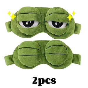 Puede incluir: Dos antifaces verdes con forma de cara de rana, con ojos blancos grandes y pupilas negras. Las m&aacute;scaras tienen una textura suave y afelpada y detalles de estrellas amarillas. El texto "2pcs" est&aacute; impreso en la parte inferior.