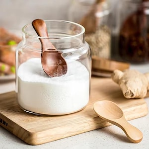 May include: A clear glass jar containing white powder with a wooden spoon inside. A small wooden spoon sits on a wooden chopping board. The jar is on a wooden surface, with other jars and spices in the background.