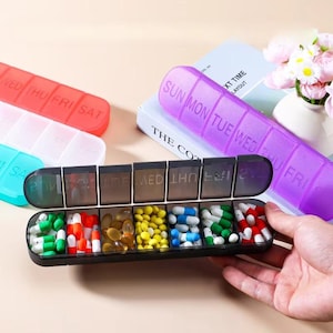 May include: A black pill organizer with seven compartments, each labeled with a day of the week. The organizer is filled with various pills in different colors. Other organizers in red, blue, and purple are also visible.