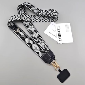 May include: Black and white patterned phone lanyard with a gold-colored clasp and a black phone holder. The lanyard features a geometric diamond design. A white card with the word "BIRTHDAY" is also visible.