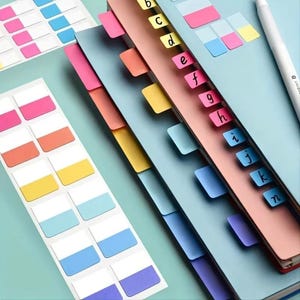 May include: An assortment of colorful sticky notes and index tabs. The image shows a notebook with lettered tabs and several sheets of rectangular sticky notes in various colors, including pink, orange, yellow, and blue. A white pen is also visible.