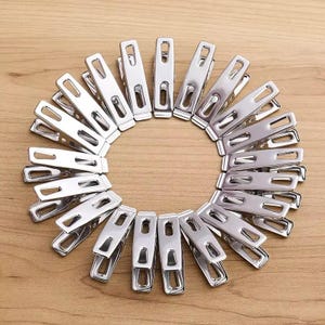 May include: A circular arrangement of shiny, silver metal clothespins. Each clothespin has a rectangular shape with a spring-loaded design. The clothespins are arranged on a light brown wooden surface.