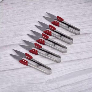 May include: Several pairs of silver-colored thread snips with red handles are arranged diagonally on a light wood-grain surface. The snips have sharp, pointed blades and a spring-loaded handle design.