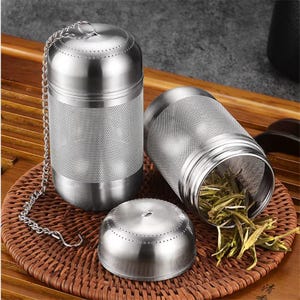 May include: Stainless steel tea infuser set. The set includes two cylindrical infusers, one with a chain, and a lid. The infusers have a perforated body and a fine mesh filter. Loose tea leaves are visible inside one of the infusers.