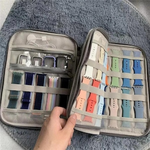 May include: Gray travel case with multiple compartments, filled with colorful watch bands in various shades. The case is open, revealing a variety of watch bands in different colors and styles, including blue, green, and pink. A hand is holding the case.