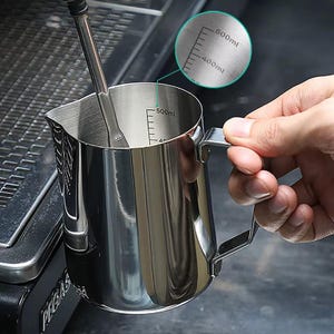 May include: A stainless steel milk frothing pitcher with a handle and internal measurement markings up to 500ml. A frothing tool is inside the pitcher. The pitcher is held by a person's hand. The background includes a coffee machine.