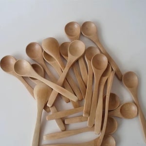 May include: A collection of small, light-colored wooden spoons. The spoons have rounded bowls and long, slender handles. The spoons are scattered across a white surface, creating a natural and rustic aesthetic.