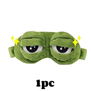 Puede incluir: Una m&aacute;scara para dormir verde con ojos grandes y de dibujos animados y una correa a juego. La m&aacute;scara tiene una textura de felpa y presenta ojos blancos con pupilas negras. Hay explosiones de estrellas amarillas a ambos lados de los ojos. El texto "1pc" est&aacute; en la parte inferior.
