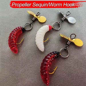 May include: Three fishing lures with the text "Propeller Sequin/Worm Hook" at the top. Each lure features a propeller, a worm-shaped body, and a hook. The worm bodies are red and white. The propellers are gold and silver.