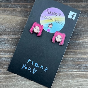 May include: A pair of pink anime-style earrings featuring a character with pink hair and a stern expression. The earrings are displayed on a black card with the text "thank you" and a logo that says "Flossy's Fashion Earrings".