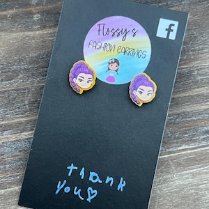 May include: A pair of stud earrings featuring a cartoon woman with purple hair and a yellow border, displayed on a black card. The card has the text "Flossy's Fashion Earrings" and "thank you" written on it.
