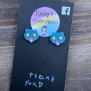 May include: A pair of blue cat-shaped earrings with yellow eyes and pink mouths are displayed on a black card. The card has the text "Flossy's Fashion Earrings" and "thank you" written in blue. A Facebook logo is visible.