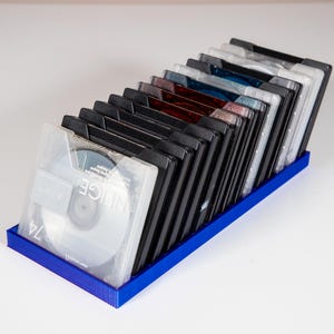 Könnte beinhalten: Ein blauer Kunststoff-Aufbewahrungskasten enthält eine Sammlung schwarzer Mini-Disc-Hüllen. Der Kasten ist mit Mini-Discs gefüllt, von denen einige mit "NICE" beschriftet sind.