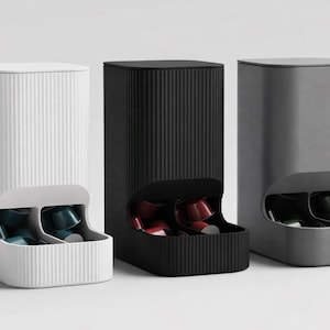 May include: Three contemporary coffee pod dispensers in white, black, and grey. Each dispenser has a rectangular form with a curved opening at the bottom, displaying the coffee pods. The white dispenser has vertical lines.