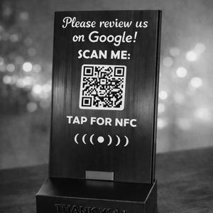 Può includere: Un cartello in bianco e nero con la scritta "Please review us on Google!" e "SCAN ME:" sopra un codice QR. Sotto il codice QR c'è "TAP FOR NFC" e una grafica. La base del cartello recita "THANK YOU!"