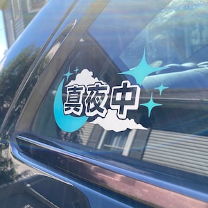 May include: A blue and white car window sticker with the Japanese text "真夜中" (pronounced "Mayanaka") which means "midnight" in English. The sticker features a crescent moon, a cloud, and sparkling stars.