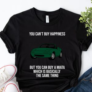 Può includere: T-shirt nero con la scritta "YOU CAN'T BUY HAPPINESS BUT YOU CAN BUY A MIATA WHICH IS BASICALLY THE SAME THING." Sotto c'è una grafica di un'auto Miata verde. La maglietta è realizzata in un materiale morbido.
